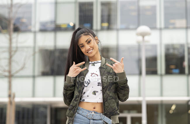Happy Asian female in trendy clothes smiling for camera and showing rock gesture on city street — Stock Photo