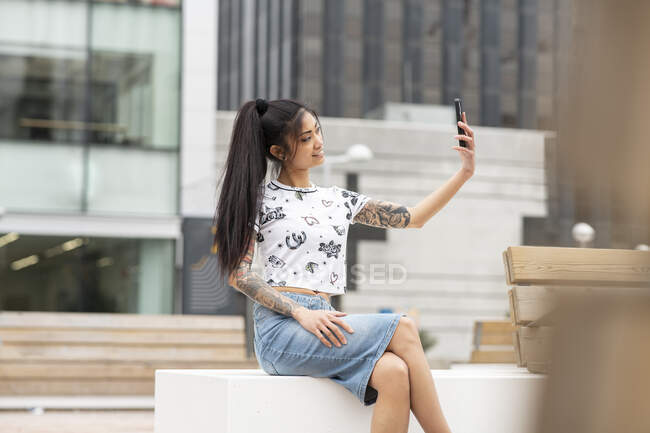 Elegante donna asiatica con braccio tatuato appoggiato sulla panchina e scattare selfie sulla strada della città — Foto stock
