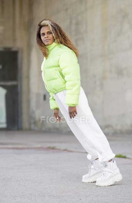 Полный вид на тело молодой афроамериканской женщины с кудрявыми волосами в стильной зеленой куртке с белыми брюками и модными сапогами, смотрящей в сторону, стоя на асфальтированной городской улице — стоковое фото