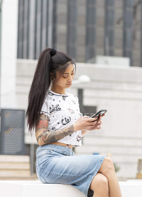 Stylish Asian female with tattooed arm resting on bench and browsing mobile phone on city street — Stock Photo