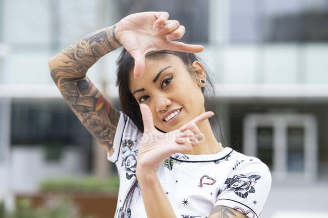 Encantada mujer asiática tatuada sonriendo y mirando a la cámara a través de un gesto de marco sobre un fondo borroso de la calle de la ciudad - foto de stock