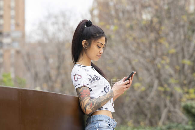 Stylish Asian female with tattooed arm leaning on bench and browsing mobile phone on city street — Stock Photo