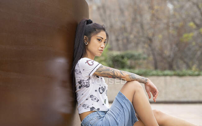Вид сбоку молодой азиатской женщины в повседневной одежде, расслабляющейся у стены и отводящей взгляд в выходные в парке — стоковое фото