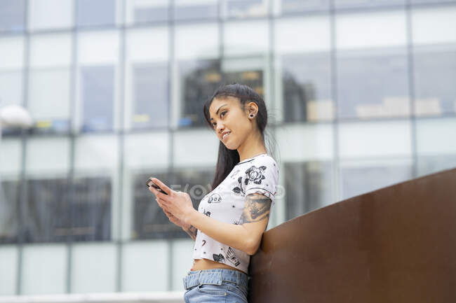 Stylish Asian female with tattooed arm leaning on wall and browsing mobile phone on city street — Stock Photo