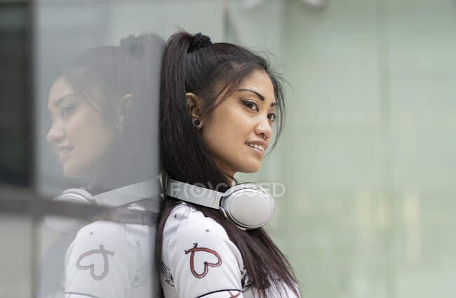 Vista lateral de la joven mujer asiática complacida con auriculares sonriendo y mirando hacia otro lado mientras se apoya en la pared de vidrio en la calle de la ciudad - foto de stock