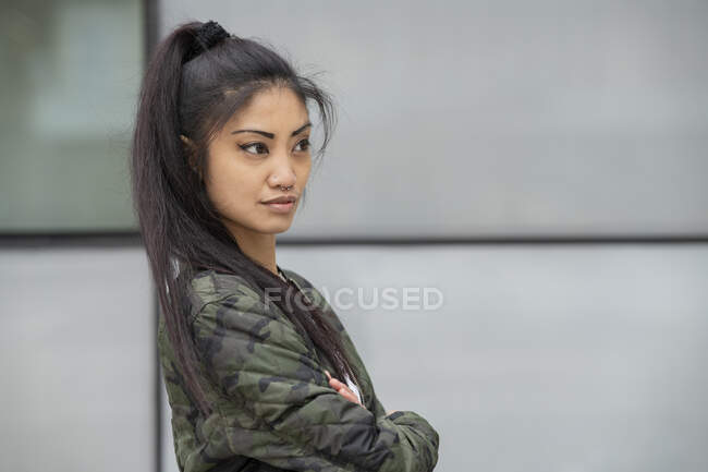 Vista lateral de la joven asiática en camuflaje de moda chaqueta cruzando brazos y mirando hacia otro lado contra la pared del edificio en la calle de la ciudad - foto de stock