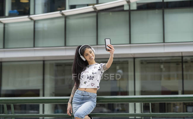 Elegante mujer asiática joven en auriculares sonriendo y tomando selfie cerca de edificio moderno en la calle de la ciudad - foto de stock