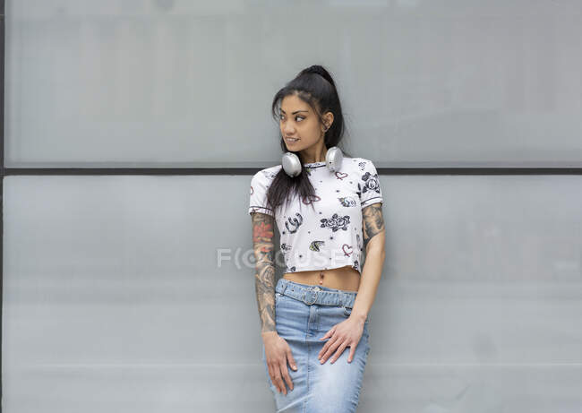 Young Asian female with headphones smiling and looking away while leaning on glass wall on city street — Stock Photo