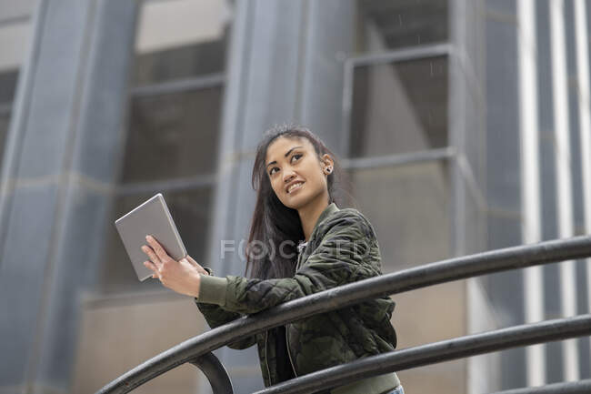 D'en bas jeune asiatique femelle en tenue décontractée navigation tablette et regarder loin avec sourire tout en s'appuyant sur rampe sur la rue de la ville — Photo de stock
