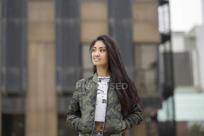 Joven mujer asiática positiva cogida de la mano en bolsillos de chaqueta de camuflaje de moda y mirando hacia otro lado con sonrisa en el fondo borroso de la calle de la ciudad - foto de stock