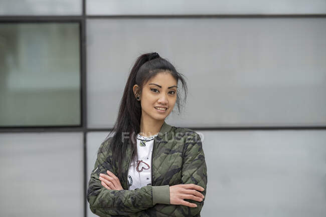 Young Asian female in trendy camouflage jacket crossing arms and looking at camera against building wall on city street — Stock Photo