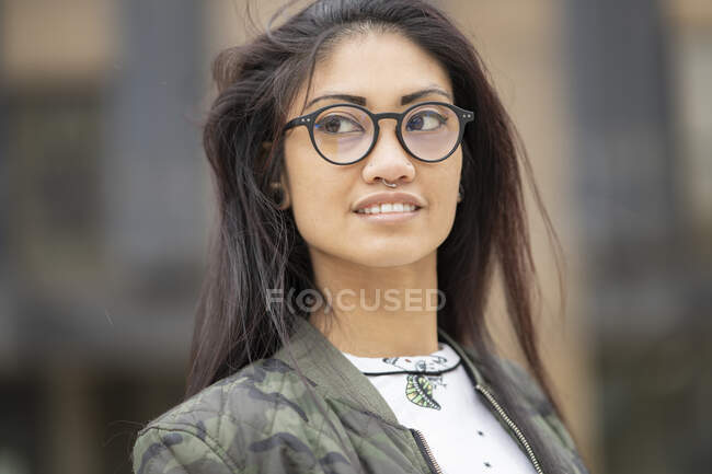 Positivo jovem Asiático fêmea na moda camuflagem jaqueta e óculos olhando para longe com sorriso no fundo borrado da rua da cidade — Fotografia de Stock