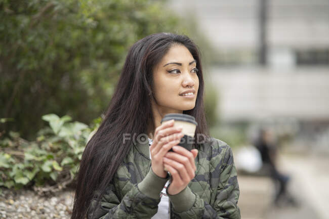 Молода азіатка в стильному одязі посміхається і озирається геть, насолоджуючись кавою, щоб піти на вихідні в парк — стокове фото