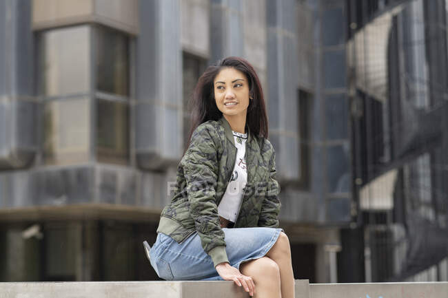 Vista lateral da jovem asiática feminina em roupas casuais relaxando perto de edifícios e olhando para longe no fim de semana — Fotografia de Stock