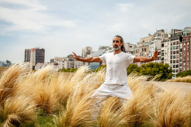 Mature bearded ethnic male in white wear stretching arms while practicing yoga and looking forward against urban buildings — Stock Photo