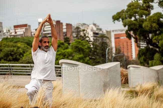 Зрілий бородатий етнічний чоловік у білому одязі розтягує руки, практикуючи йогу і з нетерпінням чекаючи міських будівель — стокове фото