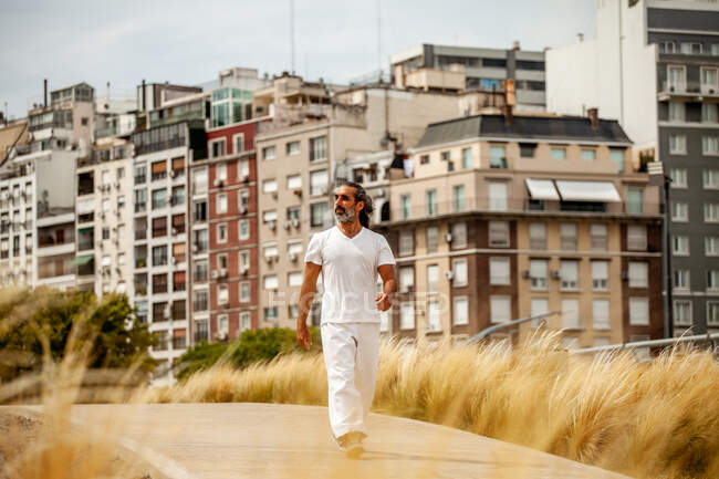 Idade média barbudo macho étnico em roupas brancas passeando na estrada e olhando para longe contra casas urbanas — Fotografia de Stock