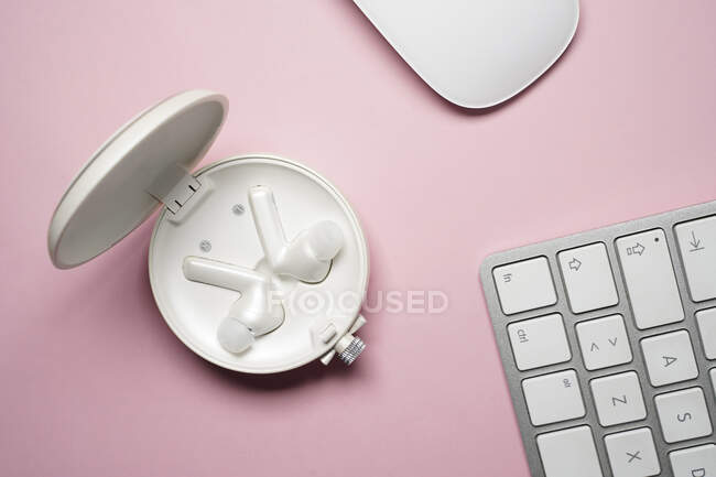 Композиция вида сверху из белых настоящих беспроводных наушников в корпусе, размещенном рядом с клавиатурой ноутбука и мышью на розовом столе — стоковое фото