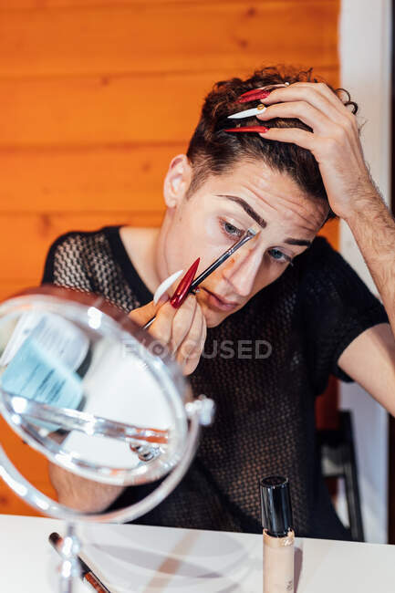 Junge fokussierte transsexuelle Männer berühren Haare beim Auftragen dekorativer Kosmetik auf Augenbrauen mit Applikator gegen Spiegel im Chalet — Stockfoto