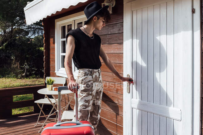 Young trendy queer with luggage opening door of wooden chalet with shadow during summer trip — Stock Photo