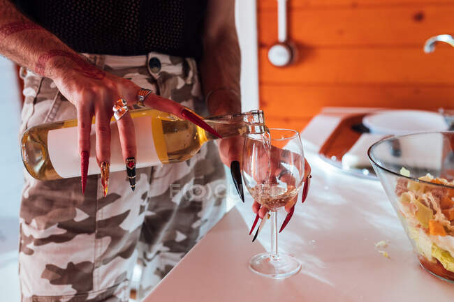 Crop homem transgênero anônimo com unhas longas que servem bebida alcoólica em vidro na mesa perto de salada de legumes em chalé — Fotografia de Stock