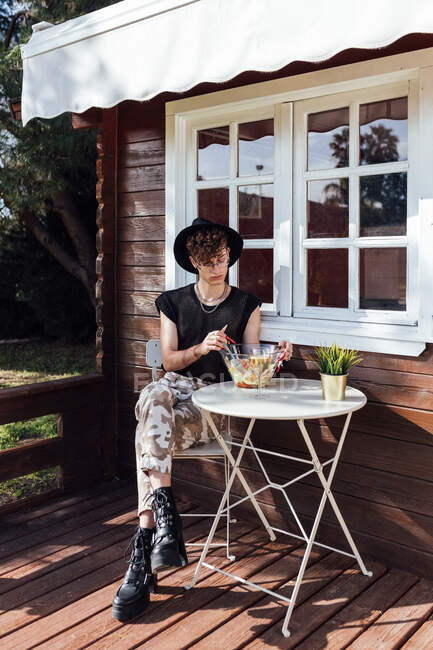 Молодой гей в модной одежде сидит со скрещенными ногами за столом с едой против бунгало — стоковое фото