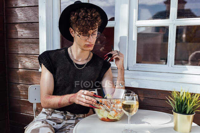 Junge Schwule in trendiger Kleidung sitzen mit gekreuzten Beinen am Tisch mit Essen gegen Bungalow — Stockfoto