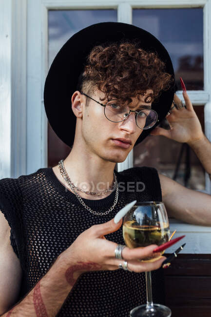Молодий транссексуал в окулярах з манікюром і келихом алкогольного напою дивиться на камеру — стокове фото