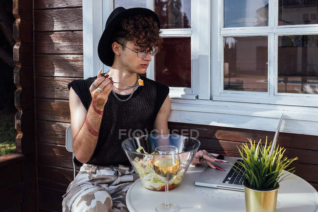 Giovane transgender uomo navigare in internet su netbook a tavola con cibo e vino bianco contro bungalow — Foto stock
