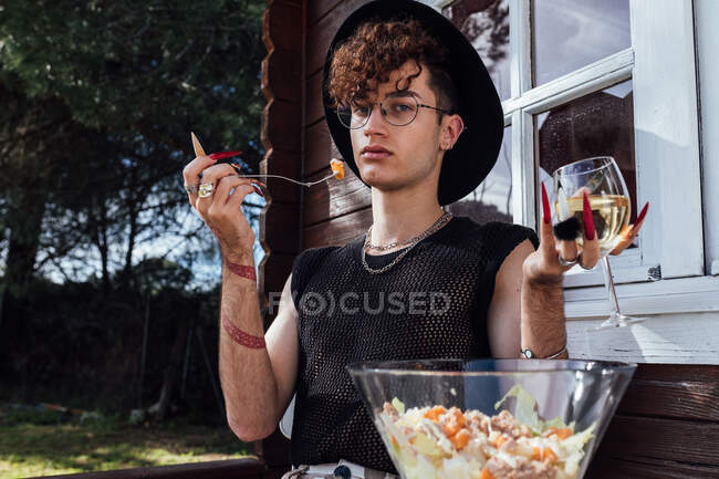 Giovane gay in moda indossare seduto a tavola con il cibo contro bungalow e vino bianco guardando la fotocamera — Foto stock