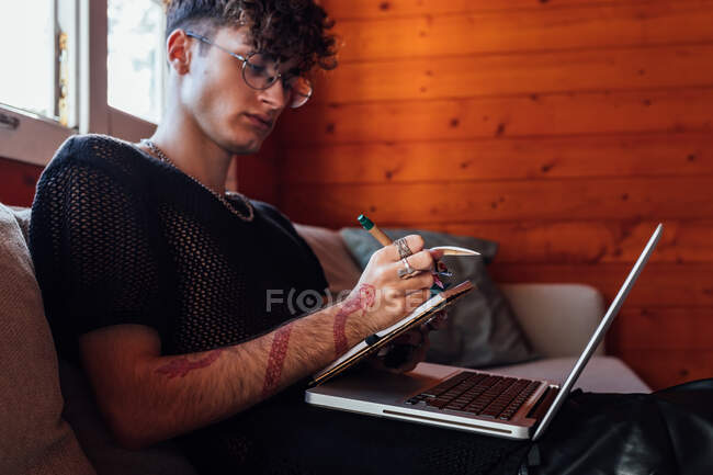 Вид збоку молодого транссексуального чоловіка, який робить нотатки в блокноті проти портативного комп'ютера, сидячи на дивані в шале — стокове фото