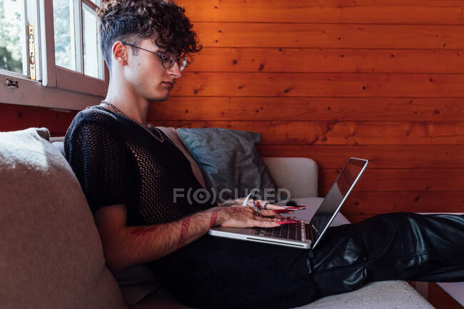 Side view of young transgender male with long nails browsing internet on netbook while resting on couch in bungalow — Stock Photo