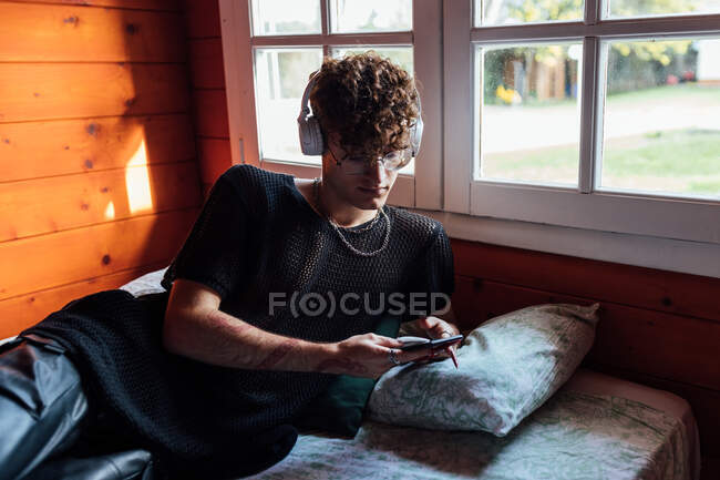 Junge Queer in drahtlosen Headset-SMS auf dem Handy, während Musik auf dem Bett in der Kabine hören — Stockfoto