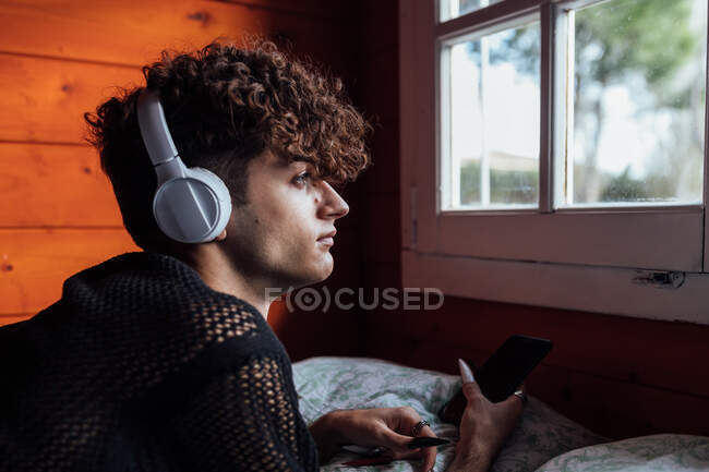 Jeune queer dans la messagerie texte casque sans fil sur téléphone portable tout en écoutant de la musique sur le lit dans la cabine — Photo de stock