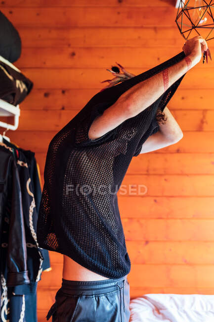 Side view of transgender male with raised arms taking off fishnet t shirt in wooden cabin — Stock Photo