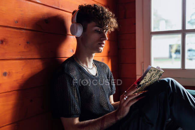 Side view of young transgender man in wireless headphones listening to song while reading diary in cabin — Stock Photo