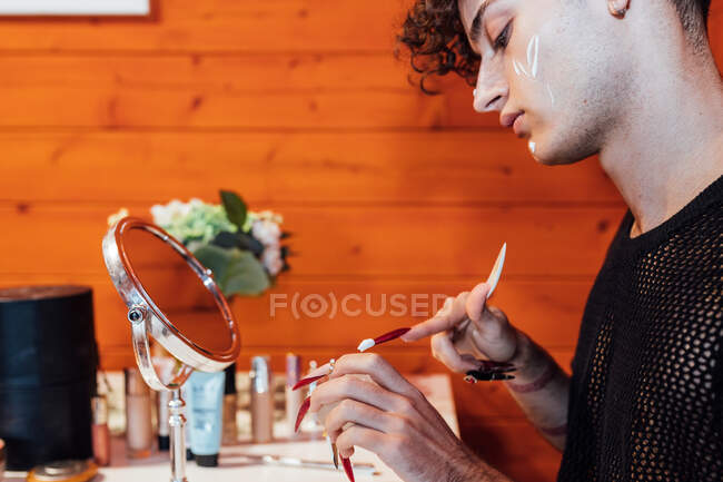 Vista lateral da colheita jovem transexual homem com creme em unhas compridas contra espelho em bangalô — Fotografia de Stock