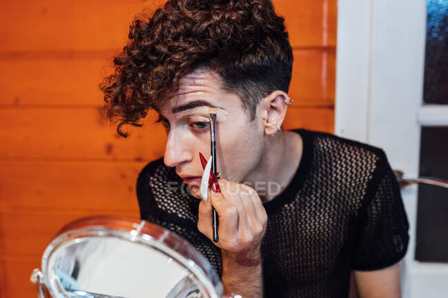 Giovane maschio transessuale focalizzato toccare i capelli mentre si applica cosmetico decorativo sul sopracciglio con applicatore contro specchio in chalet — Foto stock