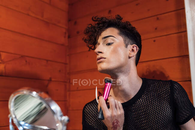 Joven transexual enfocado tocando el cabello mientras se aplica cosmética decorativa en la cara con aplicador contra espejo en chalet - foto de stock