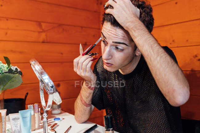 Jeune homme transsexuel concentré touchant les cheveux tout en appliquant un cosmétique décoratif sur les sourcils avec applicateur contre miroir dans le chalet — Photo de stock