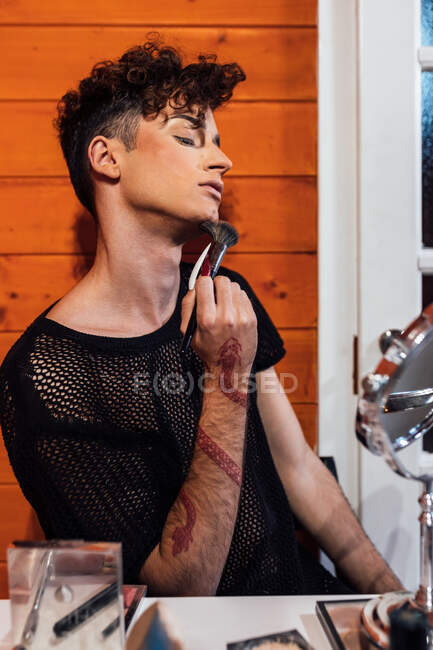 Jeune homme transsexuel avec brosse à rougir et coupe de cheveux moderne assis à table avec des produits cosmétiques dans le chalet — Photo de stock