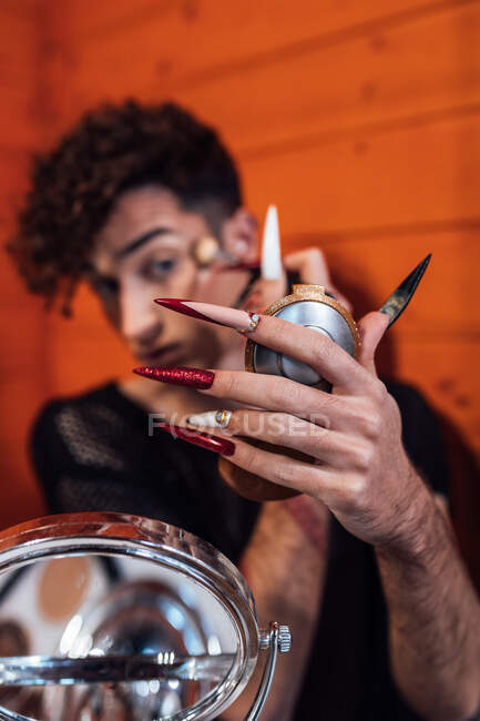 Young focused transsexual male touching hair while applying decorative cosmetic on eyebrow with applicator against mirror in chalet — Stock Photo