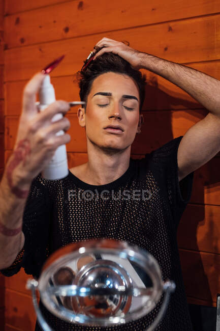 Jovem gay com fechado olhos e tatuagem pulverização cabelo a partir de garrafa perto de madeira parede no bungalow — Fotografia de Stock