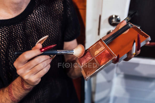 Crop queer con unghie lunghe in possesso di tavolozza rouge e pennello blush durante la preparazione per il trucco in bungalow — Foto stock