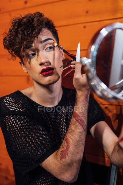 Joven transexual enfocado tocando el cabello mientras se aplica cosmética decorativa en la ceja con aplicador contra espejo en chalet - foto de stock