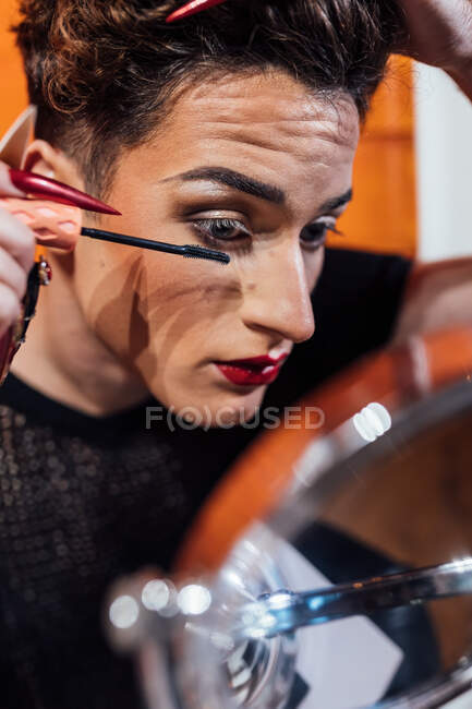 Crop young queer with manicure touching hair while applying mascara on long eyelashes against mirror — Stock Photo