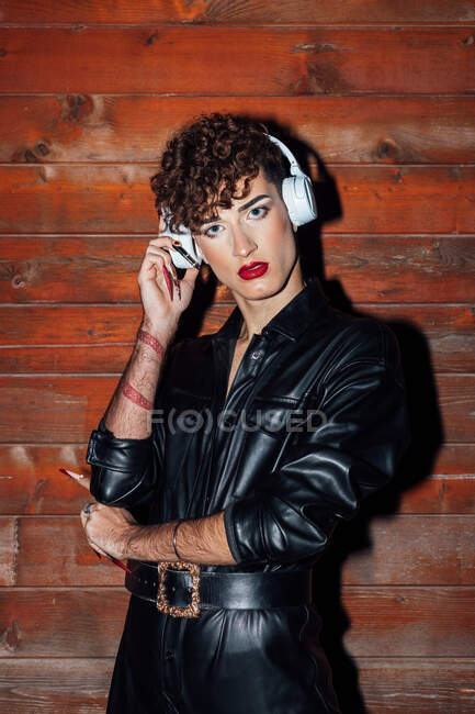 Молодой трансгендер в кожаной куртке слушает музыку из беспроводной гарнитуры, глядя на камеру в бунгало — стоковое фото