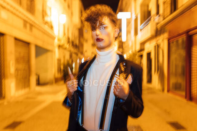 Молодой модный трансгендерный мужчина прогуливается по городской тротуаре и смотрит на камеру в сумерках — стоковое фото