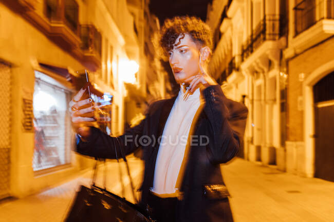 Joven hombre transgénero de moda con maquillaje tocando mejilla mientras toma autorretrato en el teléfono celular en la ciudad por la noche - foto de stock