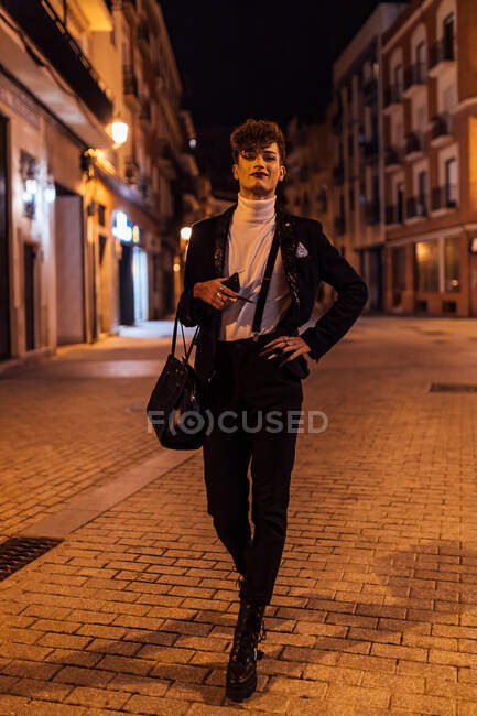 Young trendy transgender male with hand on waist strolling on urban pavement and looking at camera in twilight — Stock Photo